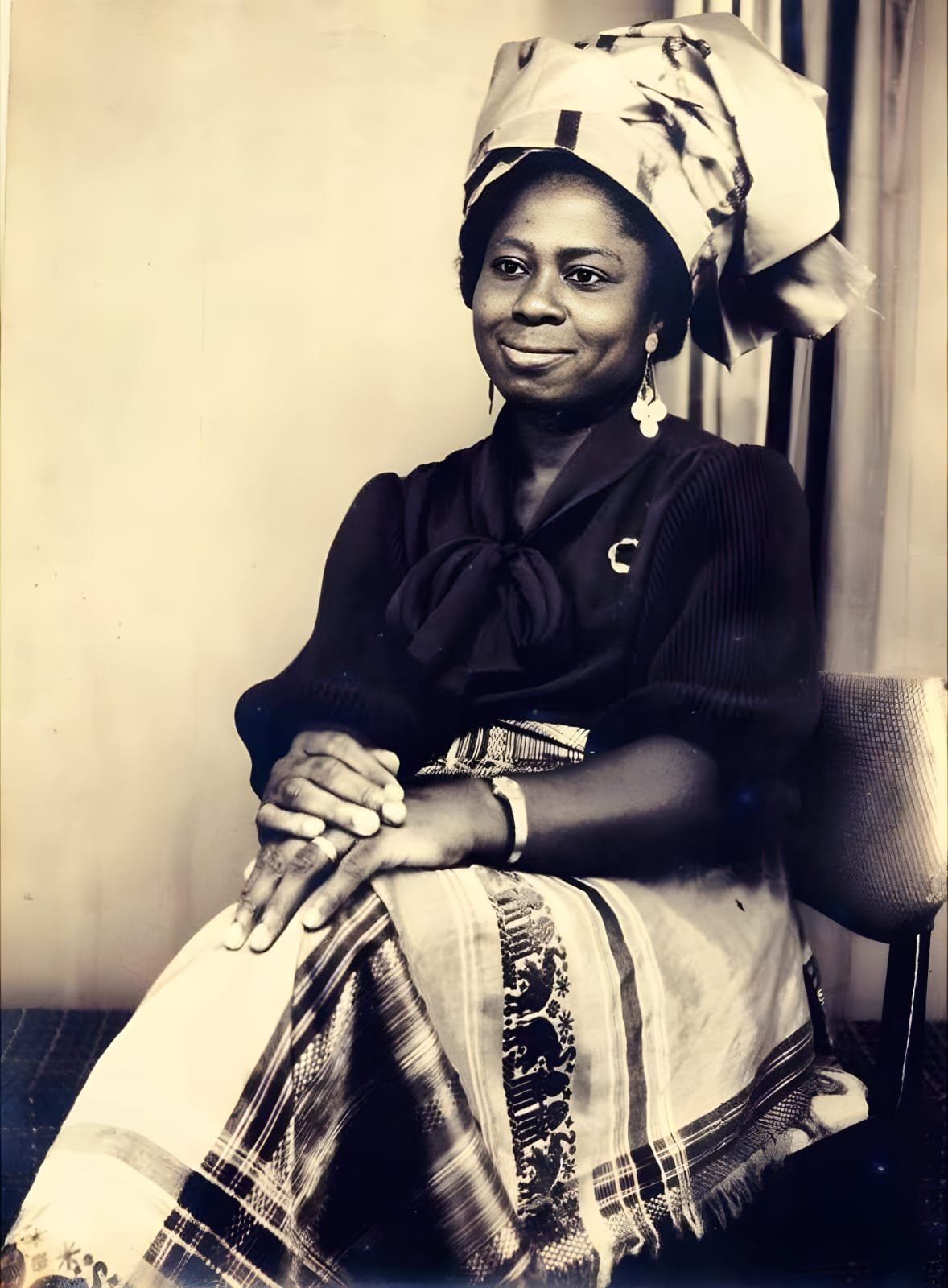 Original seated portrait of Grace Flora Oji Bakpa wearing a headwrap and patterned skirt, her hands resting in her lap